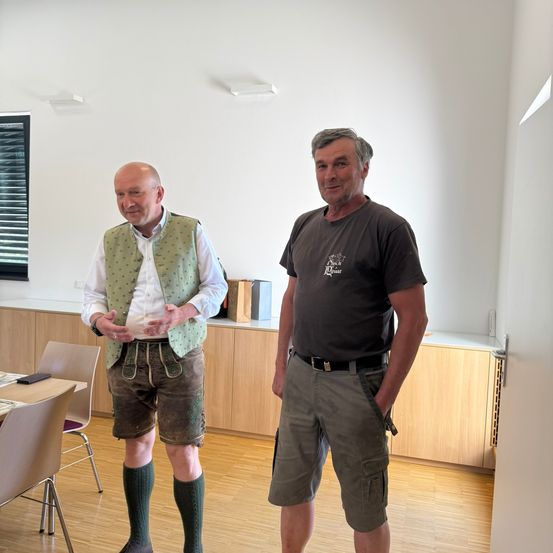 Zwei Männer stehen in einem Raum. Einer trägt eine grüne Weste und Shorts, der andere ein graues T-Shirt und Cargo-Shorts. Sie lächeln und schauen in die Kamera. Ein Tisch und Stühle sind in der Nähe.