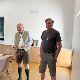 Zwei Männer stehen in einem Raum. Einer trägt eine grüne Weste und Shorts, der andere ein graues T-Shirt und Cargo-Shorts. Sie lächeln und schauen in die Kamera. Ein Tisch und Stühle sind in der Nähe.