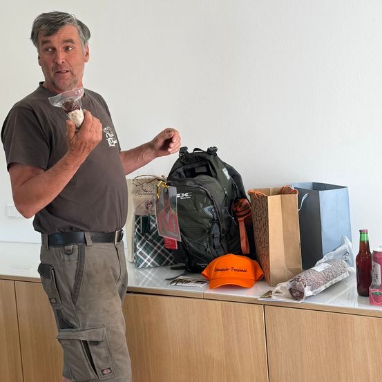 Ein Mann hält ein verpacktes Lebensmittel in Plastik und steht neben einem Tisch mit verschiedenen Gegenständen, darunter ein Rucksack, ein Papierbeutel, eine Kappe und eine Flasche.