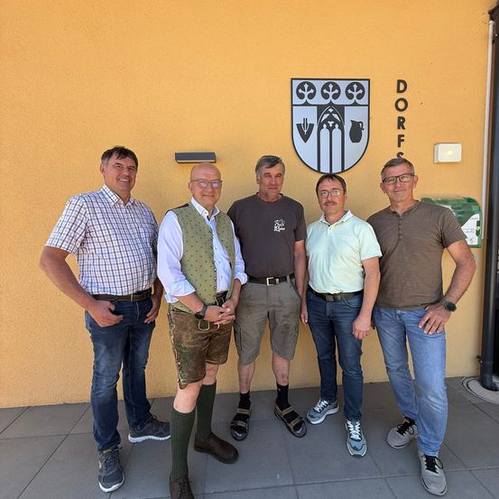 Fünf Männer stehen lächelnd vor einem gelben Gebäude. Über ihnen befindet sich ein Logo. Der Mann auf der linken Seite trägt ein kariertes Hemd.