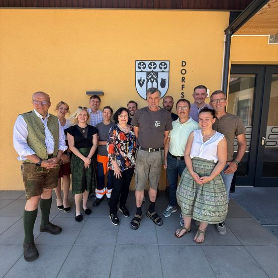 Eine Gruppe von Menschen, einige in traditioneller Kleidung, steht vor einem gelben Gebäude mit einem Logo über dem Eingang.