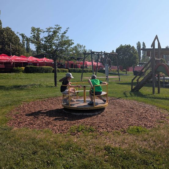 Zwei Kinder spielen auf einem Karussell in einem Park, umgeben von roten Sonnenschirmen und Bäumen. Eine Rutsche ist in der Nähe.