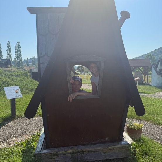Zwei Kinder schauen aus einem kleinen Holzhaus in einem Garten. Einer trägt einen Hut und der andere lächelt. Ein Schild steht in der Nähe.