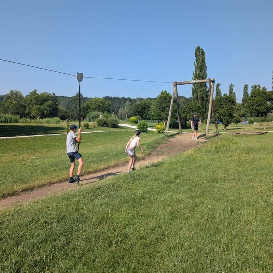 Drei Personen spielen auf einer Tauschaukel in einem Park, umgeben von Bäumen und Gras. Eine Person steht am Schaukel, während eine andere darauf zuläuft.