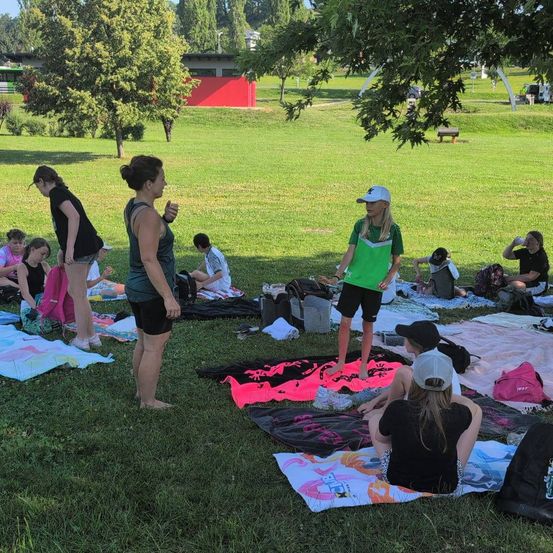 Mehrere Personen sind in einem Park versammelt. Einige sitzen auf dem Gras, während andere stehen. Eine Frau in einem grün-weißen Oberteil steht auf einer Decke. Viele Taschen und Handtücher liegen auf dem Boden.