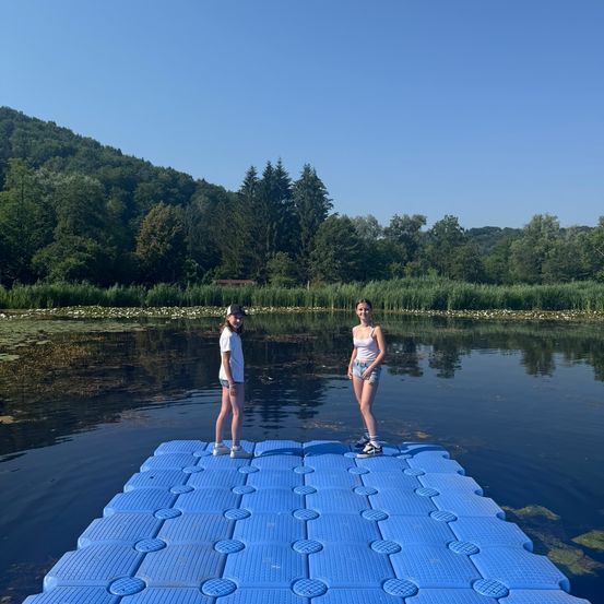 Zwei junge Frauen stehen an einem sonnigen Tag auf einer blauen schwimmenden Plattform in einem ruhigen See, umgeben von Bäumen und Wasserpflanzen.