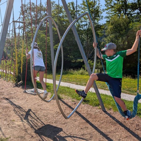 Zwei Kinder spielen auf kreisförmigen Schaukeln in einem Spielplatz. Ein Junge schwingt an einem großen Ring, während ein Mädchen an einem kleineren Ring schwingt.