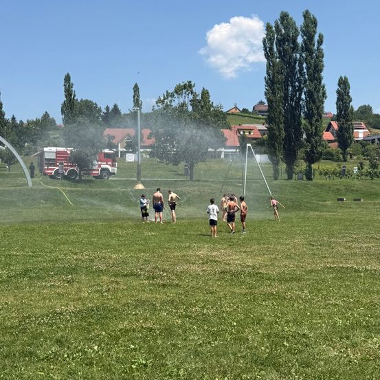 Kinder spielen an einem sonnigen Tag in einem Park mit einem Wasserwerfer, in der Nähe parkt ein Feuerwehrauto.