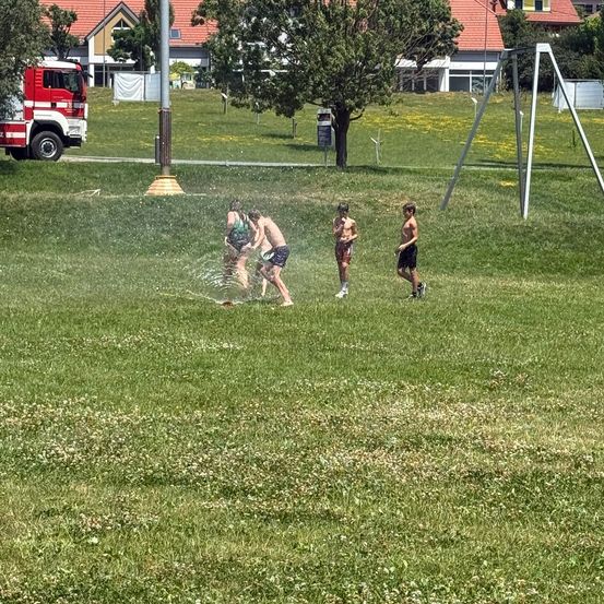 Vier junge Jungen spielen im Wasser eines Parks, einer in einem grünen Shirt spritzt den anderen mit Wasser. Dahinter steht ein Feuerwehrwagen auf der Straße.