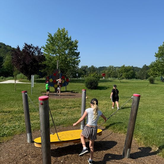 Kinder spielen auf einer gelben Wippe in einem Park mit einer bunten Kletterstruktur, Bäumen und blauem Himmel.
