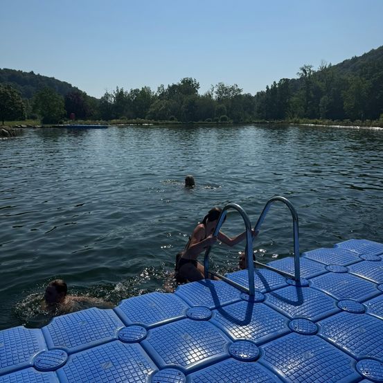 Ein ruhiger See mit schwimmenden Menschen, einer nahe dem blauen Steg. Bäume und Berge säumen den Seerand unter einem klaren Himmel.