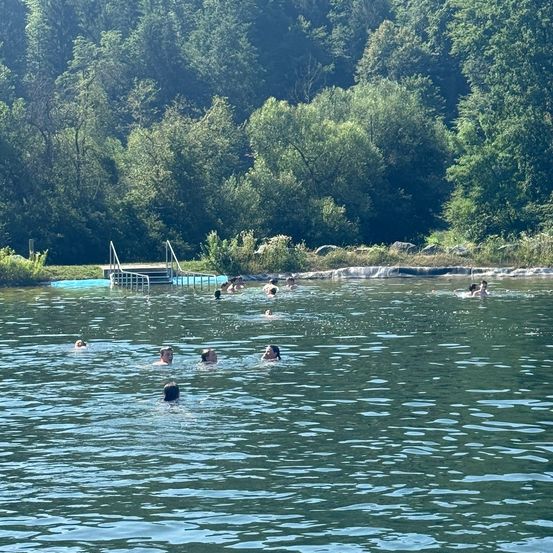 Eine Gruppe von Menschen schwimmt in einem See unter einem hellen Himmel mit einer Holzbrücke im Hintergrund.