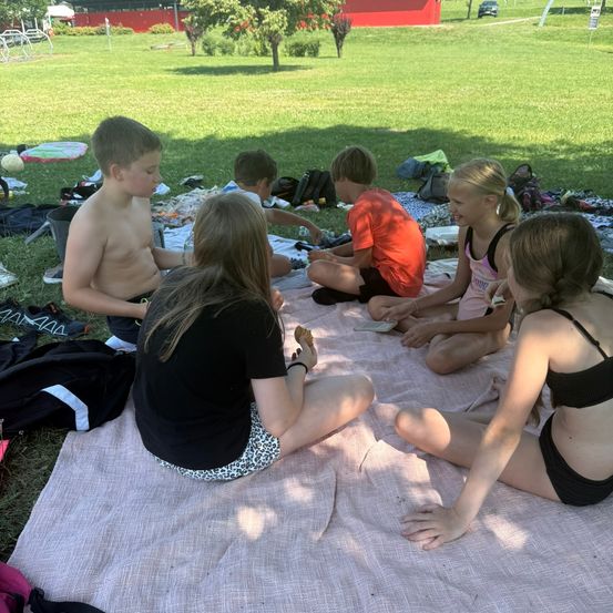 Eine Gruppe von Kindern in Badekleidung sitzt auf einer Decke in einem Park. Sie sind von Taschen und Spielzeug umgeben. Ein Junge ohne Hemd schaut eine Mädchen an.