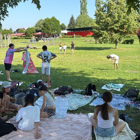 Eine Gruppe von Menschen genießt einen sonnigen Tag im Park. Einige sitzen auf Decken, andere stehen. Eine Person in einem grauen Shirt beugt sich. In der Ferne geht eine Frau mit einem Kind.