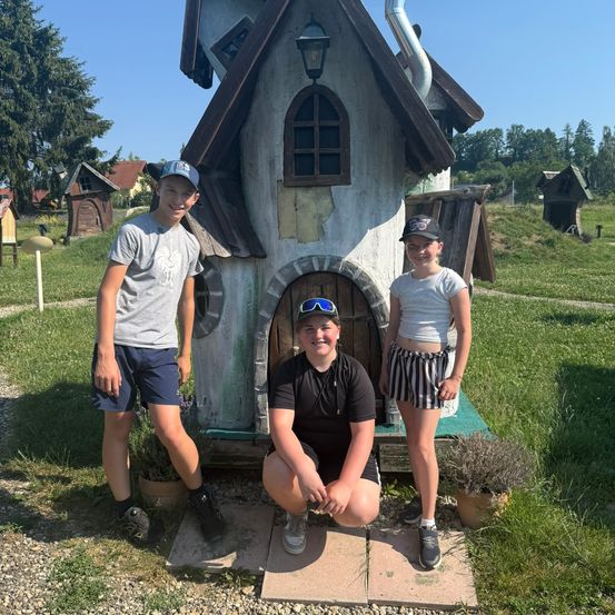 Drei junge Personen posieren für ein Foto vor einem verspielten Haus. Ein Junge kniet mit einer Frau auf seinem Schoß, während ein anderer Junge neben ihnen steht.