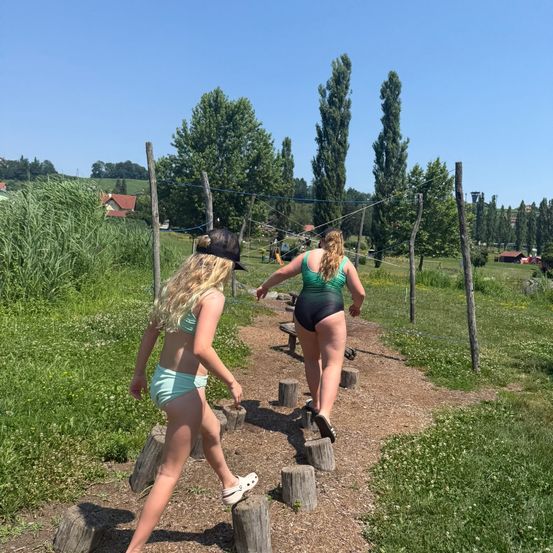 Zwei Frauen in Badekleidung gehen auf einem Pfad mit Trittsteinen. Sie navigieren durch einen Rasenbereich mit Bäumen und einem Volleyballnetz im Hintergrund.