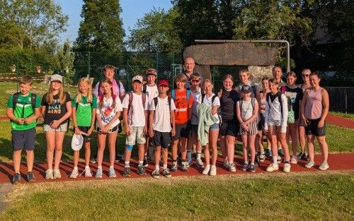 Eine Gruppe von Kindern und Erwachsenen steht an einem sonnigen Tag im Freien, trägt Freizeitkleidung und Turnschuhe. Sie stehen vor einem Grasbereich mit Bäumen und einer Steinstruktur.