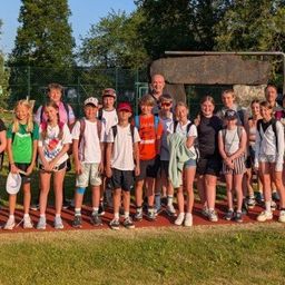Eine Gruppe von Kindern und Erwachsenen steht an einem sonnigen Tag im Freien, trägt Freizeitkleidung und Turnschuhe. Sie stehen vor einem Grasbereich mit Bäumen und einer Steinstruktur.