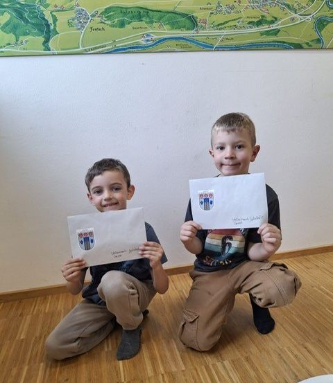 Zwei junge Jungen knien und halten Urkunden hoch, wahrscheinlich in einem Klassenzimmer. Beide Jungen lächeln und sehen aufgeregt aus. Hinter ihnen hängt eine Karte an der Wand.
