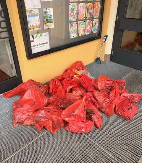 Eine Ansammlung roter Plastiktüten, möglicherweise mit Abfall, ist auf einem Metallrost vor einem Gebäude aufgetürmt. Papiere sind am Anschlagbrett befestigt.