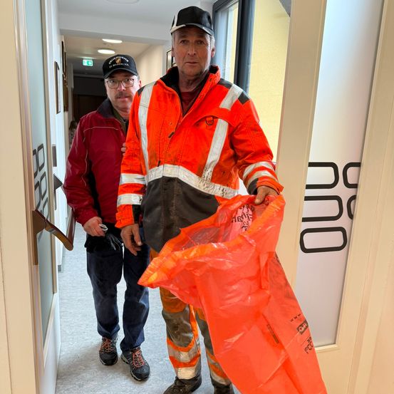Zwei Männer stehen in einem Korridor. Einer hält eine Plastiktüte. Beide tragen reflektierende Kleidung. Der Mann links trägt eine Brille und einen Hut.