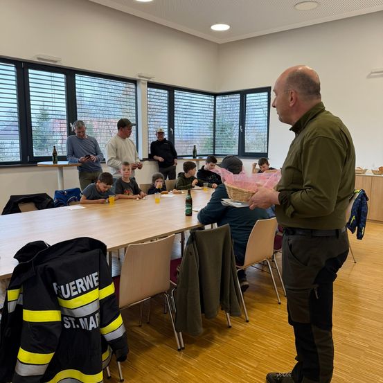 Eine Gruppe von Menschen, darunter Kinder, sitzt um einen Tisch in einem Konferenzraum herum. Ein Mann in einem grünen Shirt hält einen Korb.