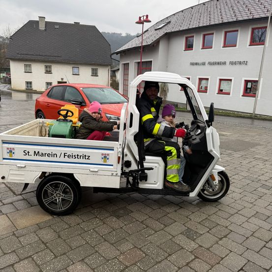 Ein Feuerwehrauto mit einem Feuerwehrmann und zwei Kindern fährt auf einer gepflasterten Straße mit einem roten Auto im Hintergrund.