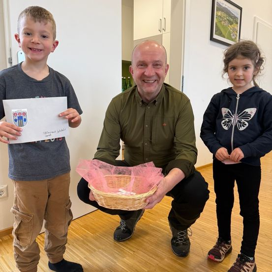 Ein Mann lächelt, während er sich bückt und einen Korb hält, mit zwei Kindern neben ihm. Der Junge hält ein Zertifikat.