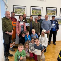 Eine Gruppe von Erwachsenen und Kindern posiert für ein Foto in einem Raum mit Holzboden und gerahmten Bildern an der Wand. Einige halten Urkunden in den Händen, andere lächeln in die Kamera.