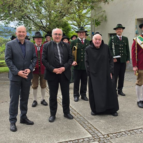 Eine Gruppe von Männern in traditioneller deutscher Kleidung, darunter ein Mann in einem schwarzen Gewand, steht vor einem Gebäude. Einige tragen Musikinstrumente.