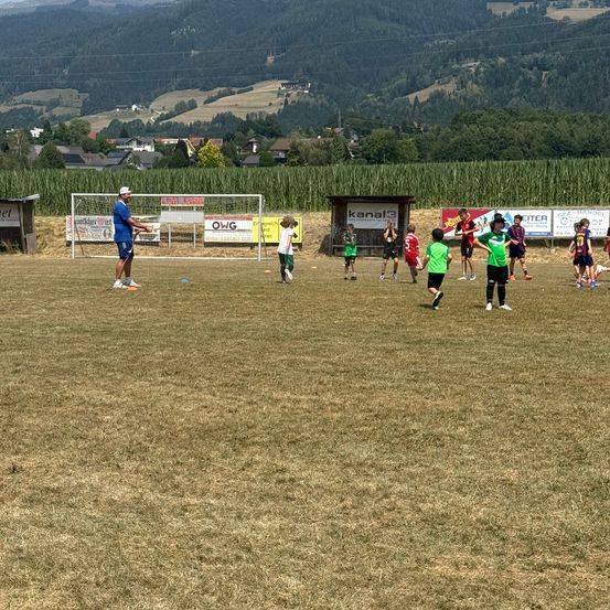 Eine Gruppe von Kindern in grünen und roten Trikots spielt Fußball auf einem Feld mit einem Trainer in Blau. Das Feld ist von einem Maisfeld umgeben und im Hintergrund befinden sich Berge.