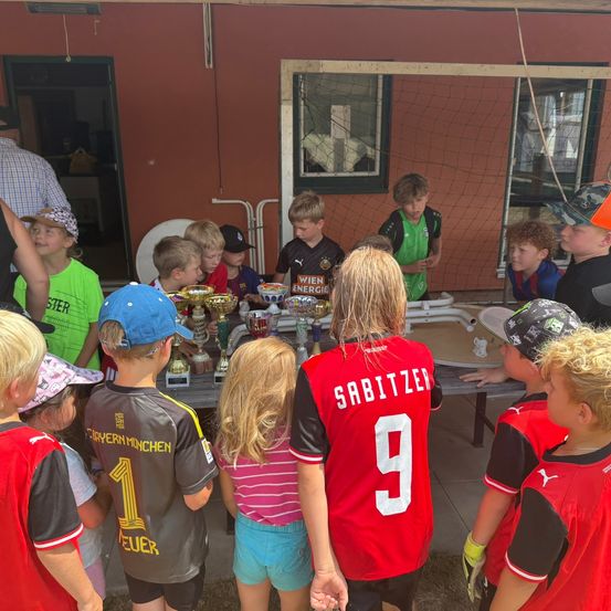 Eine Gruppe von Kindern in Sporttrikots versammelt sich um einen Tisch mit Trophäen. Einige tragen Mützen, und ein Kind in einem roten Trikot mit der Nummer 9 steht vorne. Dahinter ist ein Gebäude mit einem Fenster und einem Netz zu sehen.
