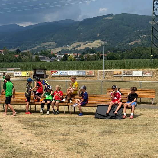 Eine Gruppe junger Jungen in Sportkleidung sitzt auf einer Bank auf einem Feld, mit Bergen und einem bewölkten Himmel im Hintergrund. Einige von ihnen tragen Helme und Turnschuhe.