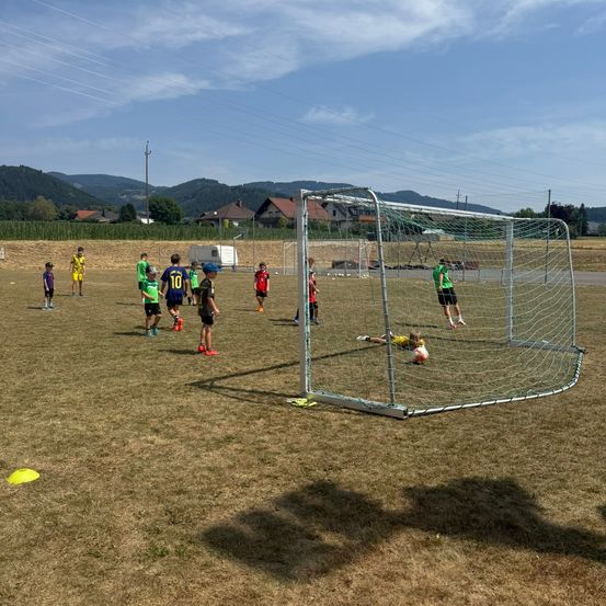 Eine Gruppe junger Fußballspieler trainiert auf einem Feld mit einem Tornetz im Vordergrund. Die Spieler tragen verschiedenfarbige Trikots.