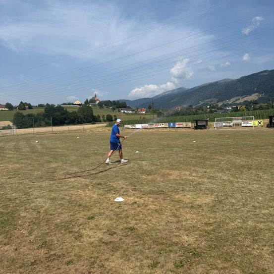 Ein Mann in einem blauen Shirt und Shorts trainiert mit einer Schnur auf einem Grasfeld mit einer Kulisse aus Bergen und Häusern.