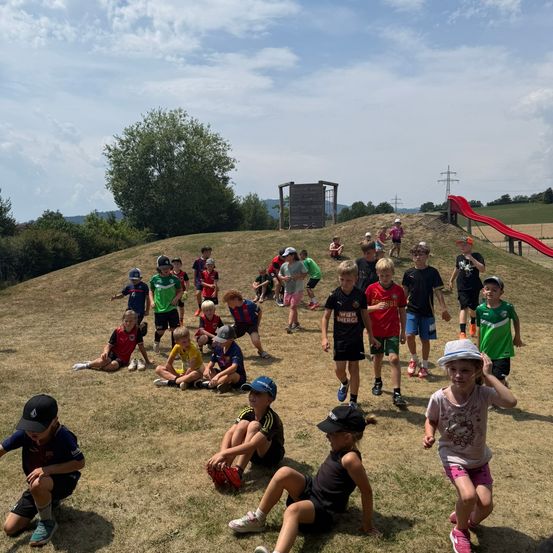 Eine Gruppe von Kindern in Sportkleidung spielt auf einem Grashügel mit einer Rutsche im Hintergrund. Einige sitzen, andere stehen und einige gehen.