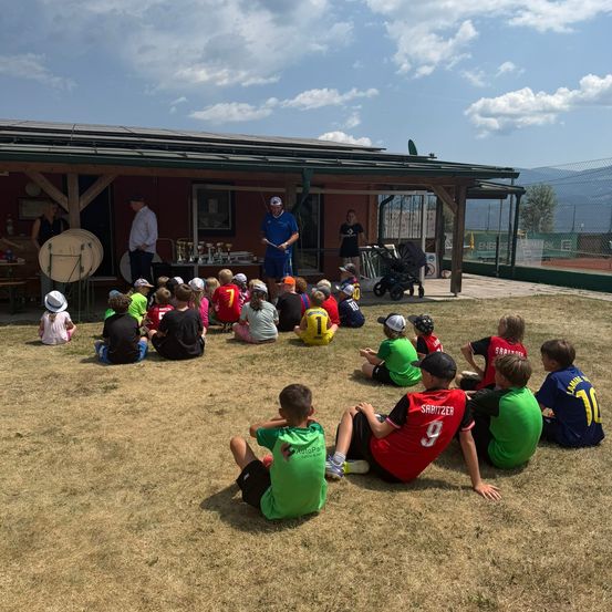 Eine Gruppe von Kindern in Sporttrikots sitzt im Kreis auf einem Feld, vor ihnen stehen zwei Erwachsene vor einem Gebäude. Einer der Erwachsenen hält einen Schläger, der andere steht in der Nähe eines Kinderwagens.