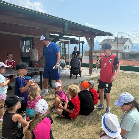 Eine Gruppe von Kindern und Erwachsenen versammelt sich auf einem Rasenfeld. Ein Mann in einem blauen Shirt steht vor ihnen und hält eine Trophäe. Die Kinder sitzen auf dem Boden, einige tragen Hüte und Turnschuhe. Hinter ihnen befindet sich ein Gebäude mit einem Zaun und einem Kinderwagen. In der Ferne sind Häuser und Berge zu sehen.