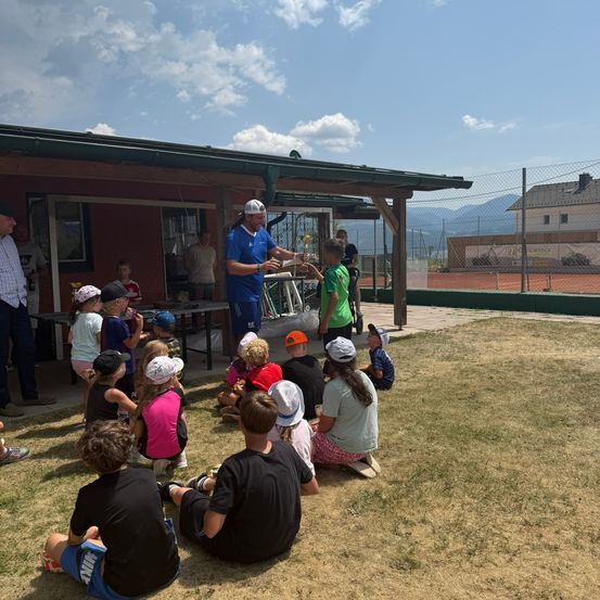 Eine Gruppe von Kindern und Erwachsenen versammelt sich um einen Mann, der einen Pokal und einen Tennisschläger hält, wahrscheinlich in einer Sportanlage.