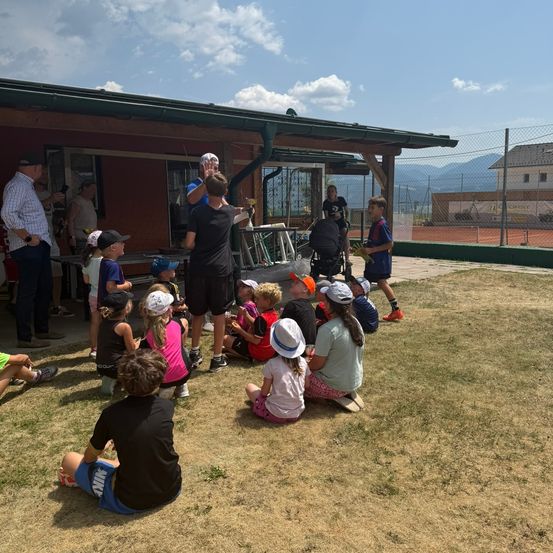 Eine Gruppe von Kindern und Erwachsenen versammelt sich auf einer Rasenfläche, möglicherweise für eine Sportveranstaltung. Einige sitzen, andere stehen, und einige tragen Hüte. Ein Gebäude mit Zaun und Tennisplatz ist im Hintergrund zu sehen.