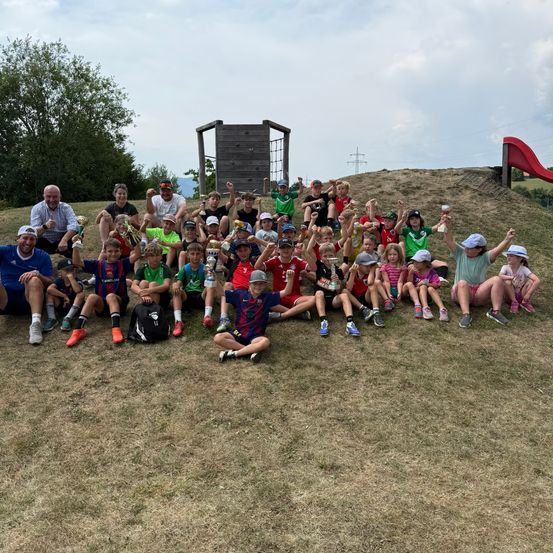 Eine Gruppe von Kindern und Erwachsenen posiert auf einem Grashügel für ein Foto und hält Pokale in den Händen.