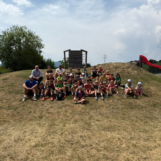 Eine Gruppe von Kindern und Erwachsenen sitzt auf einem Grashügel und posiert für ein Foto mit einer roten Rutsche im Hintergrund.