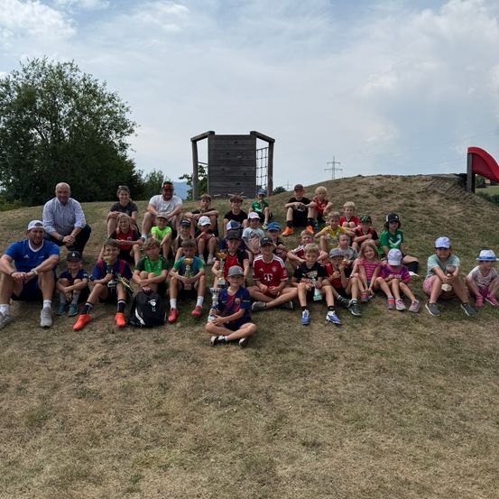Eine Gruppe von Kindern und Erwachsenen posiert für ein Foto auf einem Grashügel mit einer Holzstruktur und einer Rutsche im Hintergrund.