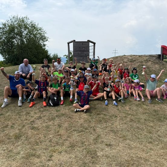 Eine Gruppe von Kindern und Erwachsenen posiert für ein Foto auf einem Grasfeld, hält Pokale und Tassen. Einige tragen Hüte und Turnschuhe. Dahinter steht eine Holzstruktur und ein Baum.