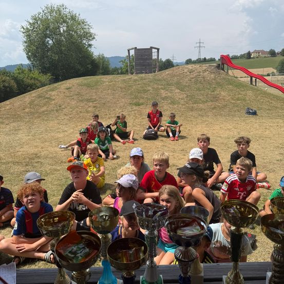 Eine Gruppe von Kindern und Jugendlichen sitzt auf dem Boden, einige tragen Mützen und halten Pokale. Sie sind von einem Grashügel umgeben, im Hintergrund befindet sich eine Rutsche.