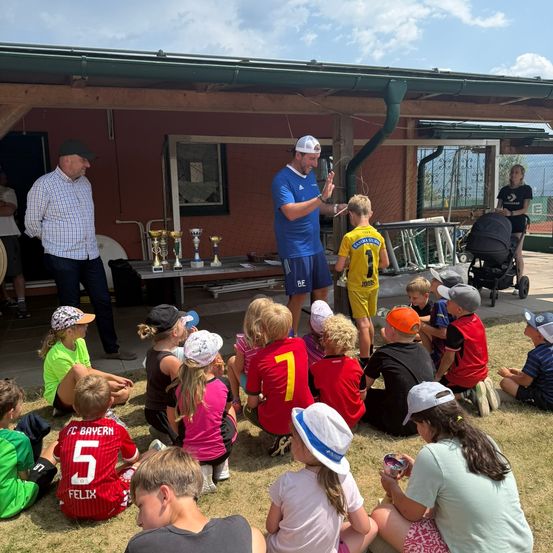 Eine Gruppe von Kindern und Erwachsenen ist um einen Mann in einem blauen Shirt versammelt, wahrscheinlich ein Trainer, der zu ihnen spricht. Sie sitzen auf dem Rasen vor einem Gebäude mit Trophäen auf einem Tisch.