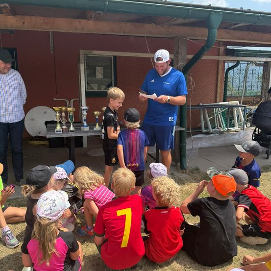 Eine Gruppe von Kindern in Sporttrikots ist um einen Mann in einem blauen Shirt und einer weißen Kappe versammelt, der von einem Stück Papier liest. Sie stehen vor einem Gebäude mit Trophäen auf einem Tisch.