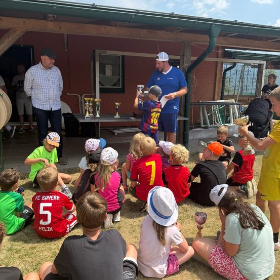 Eine Gruppe von Kindern und Erwachsenen versammelt sich vor einem Gebäude, einige halten Trophäen und Pokale. Ein Mann in einem blauen Shirt überreicht einem Kind eine Trophäe.