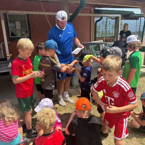 Eine Gruppe von Kindern in Sporttrikots versammelt sich um einen Mann in einem blauen Shirt, der ein Papier hält und Gegenstände zu verteilen scheint. Die Kinder schauen den Mann und einander an, einige halten Gegenstände. Hinter ihnen steht ein Gebäude mit einem Fenster und einem Zaun.