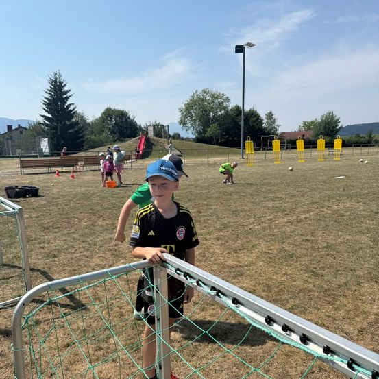Ein junger Junge in einem Fußballtrikot steht neben einem Tornetz auf einem Feld, auf dem andere Kinder Fußball spielen.